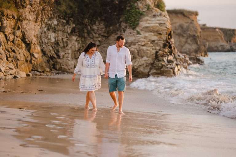 Couple laughing and walking along sandy Cornish Beach - The Perfect Engagement: Your Ultimate Guide To Plan Your Unique Proposal
