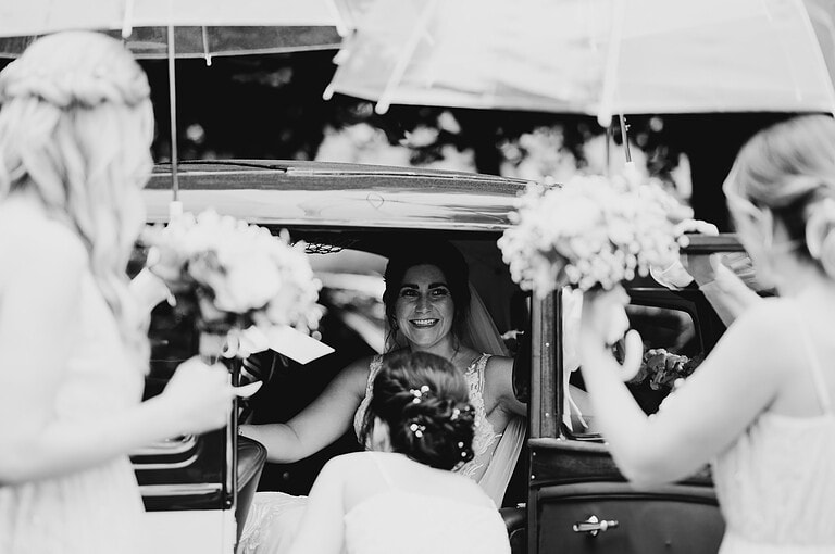 Bride getting out of her classic car under umbrellas as the rain comes down - How To Pick Your Perfect UK Wedding Venue