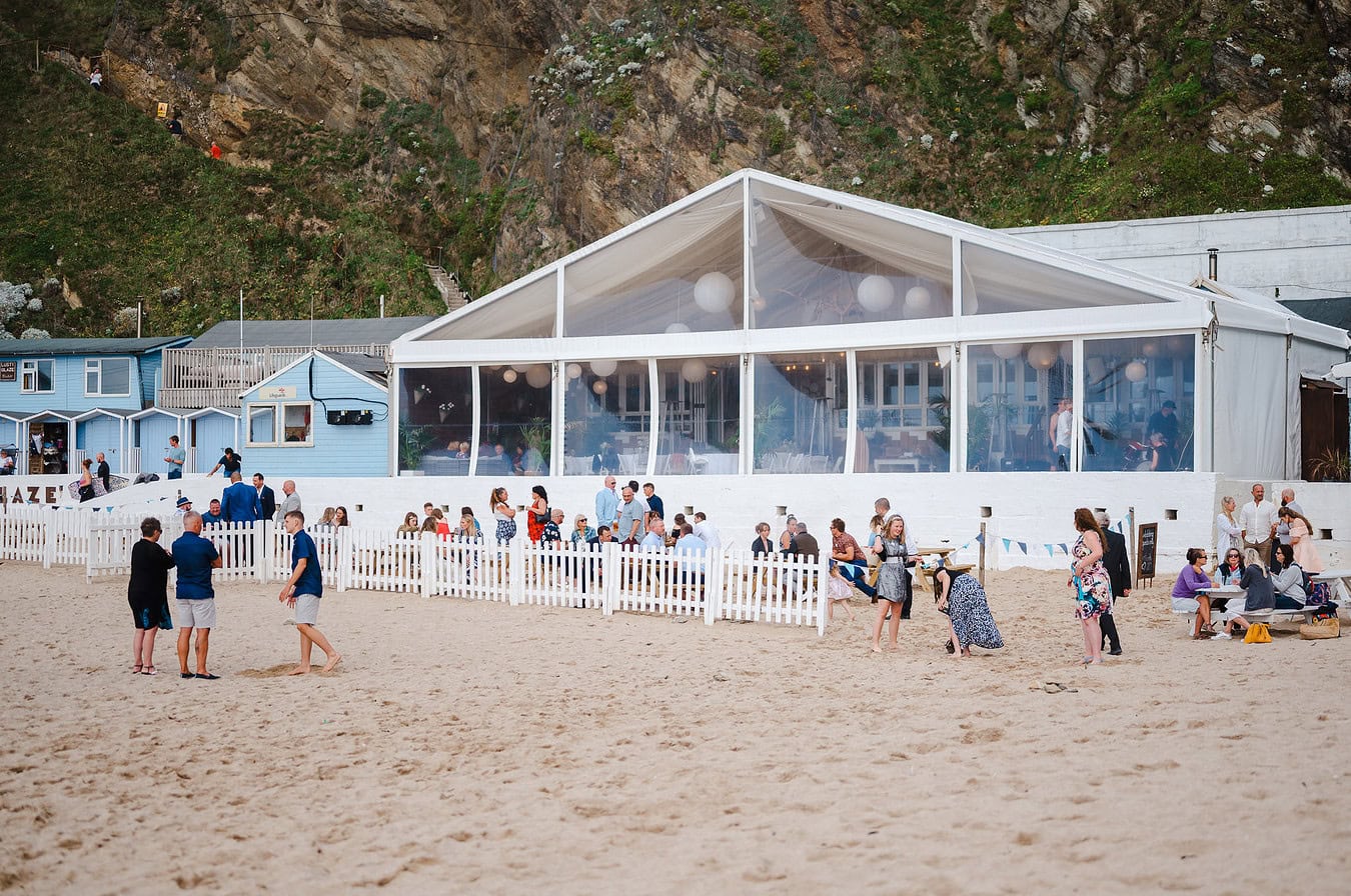 View looking back on Lusty Glaze as wedding guests hand around the beach tables