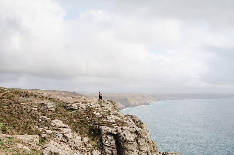 Looking across the cliffs at the couple standing on the edge of the St Agnes coast path arms wrapped around each other. By Cornwall Engagement Photographer, Jake Timms Photography