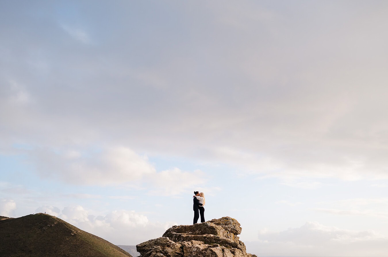 Standing on top of the boulderas the couple hug and put their arms around each other at Chapel Porth, Cornwall