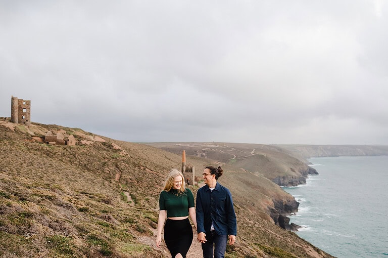 Hapily walking and laughing as they walk along the St Agnes coast path with a Cornish mine in the background as the happy couple enjoy their Chapel Porth engagement photo shoot