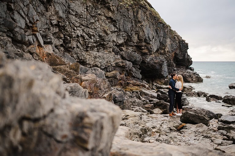 A rocky backdrop and a pebble shore, the couple perch on a boulder and kiss as the enjoy their engagement photo shoot