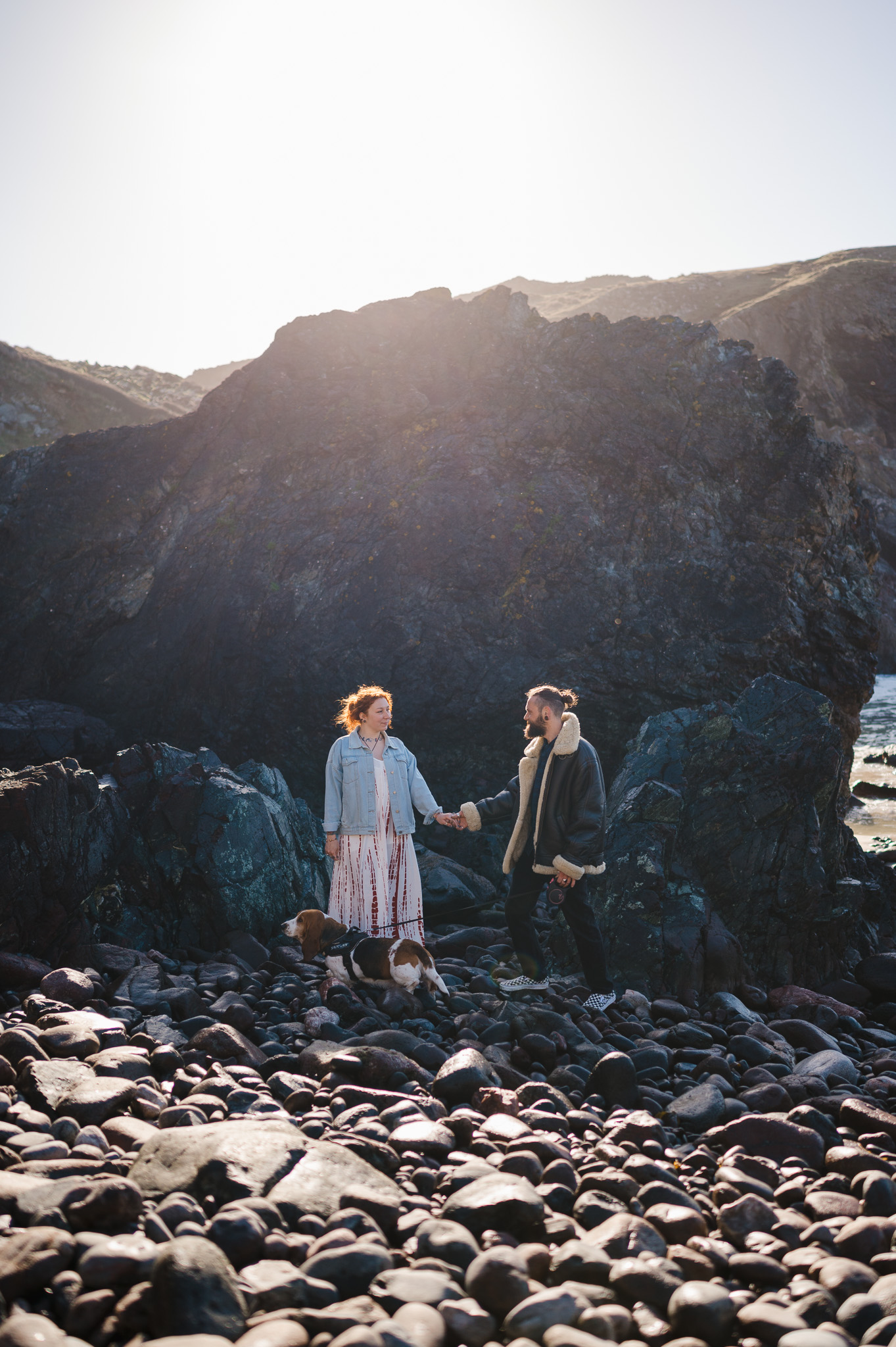 Standing in front of the large rock as the sun starts to shine over the top, taken during their engagement photo shoot at Kynance Cove