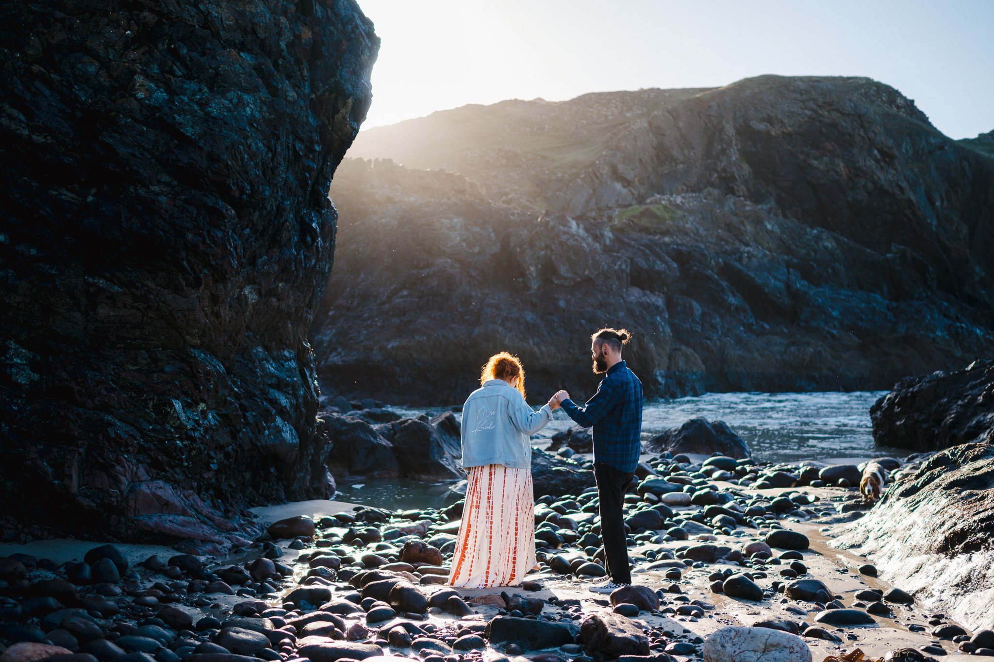 The couple help each other as they walk along the rocky shore of Kynance Cove during their Kynance Cove Engagement photo shoot