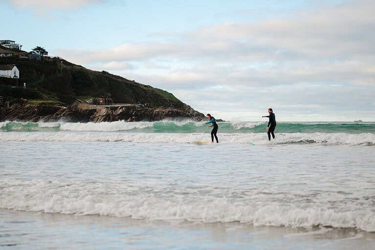 Standing up on the surfboards together to ride a wave into the shore at Towans Beach, Newquay. By Cornwall Engagement Photographer