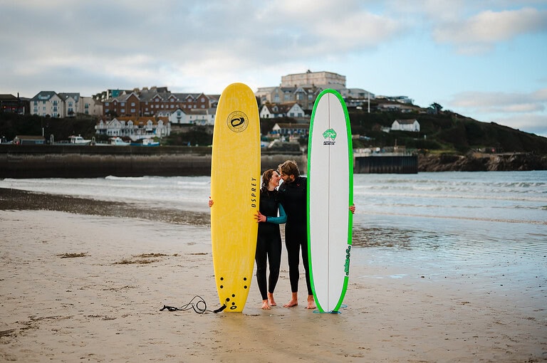 Taking a kiss whilst holding their surfboards on a beach in Cornwall by Cornwall engagement photographer