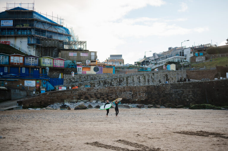 A newly engaged couple walk across Newquay beach with surfboards on their head during their Newquay Engagement Photo Shoot