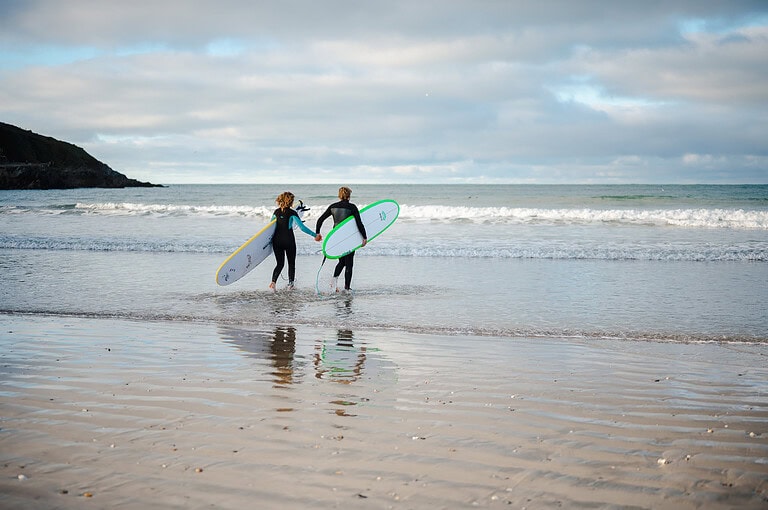 Running into the cold waters with surfboards in hand during their Newquay engagement photo shoot by Cornwall engagement photo shoot