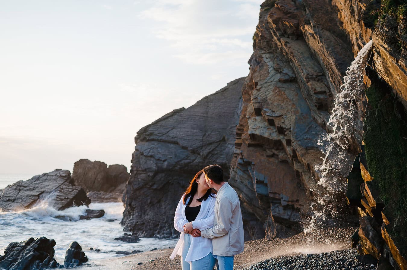 Arms wrapped around each other as the waves hit the rocky shore and as the waterfall falls behind them at sunset during their Sandymouth engagement photo shoot