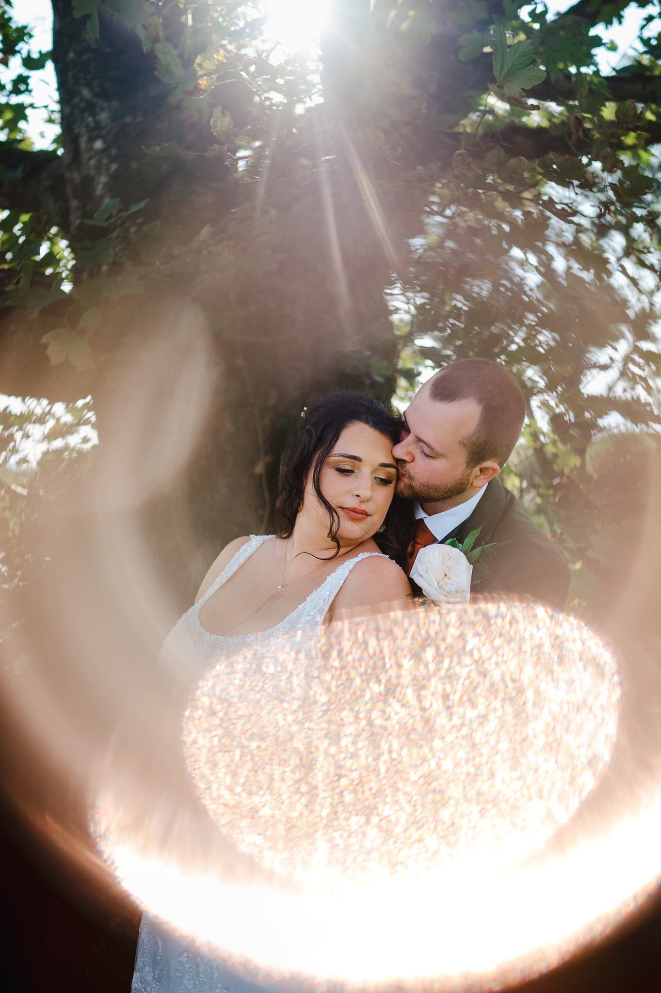 Newlyweds stand by tree as the golden light shines through from behind by London Wedding Photographer, Jake Timms Photography