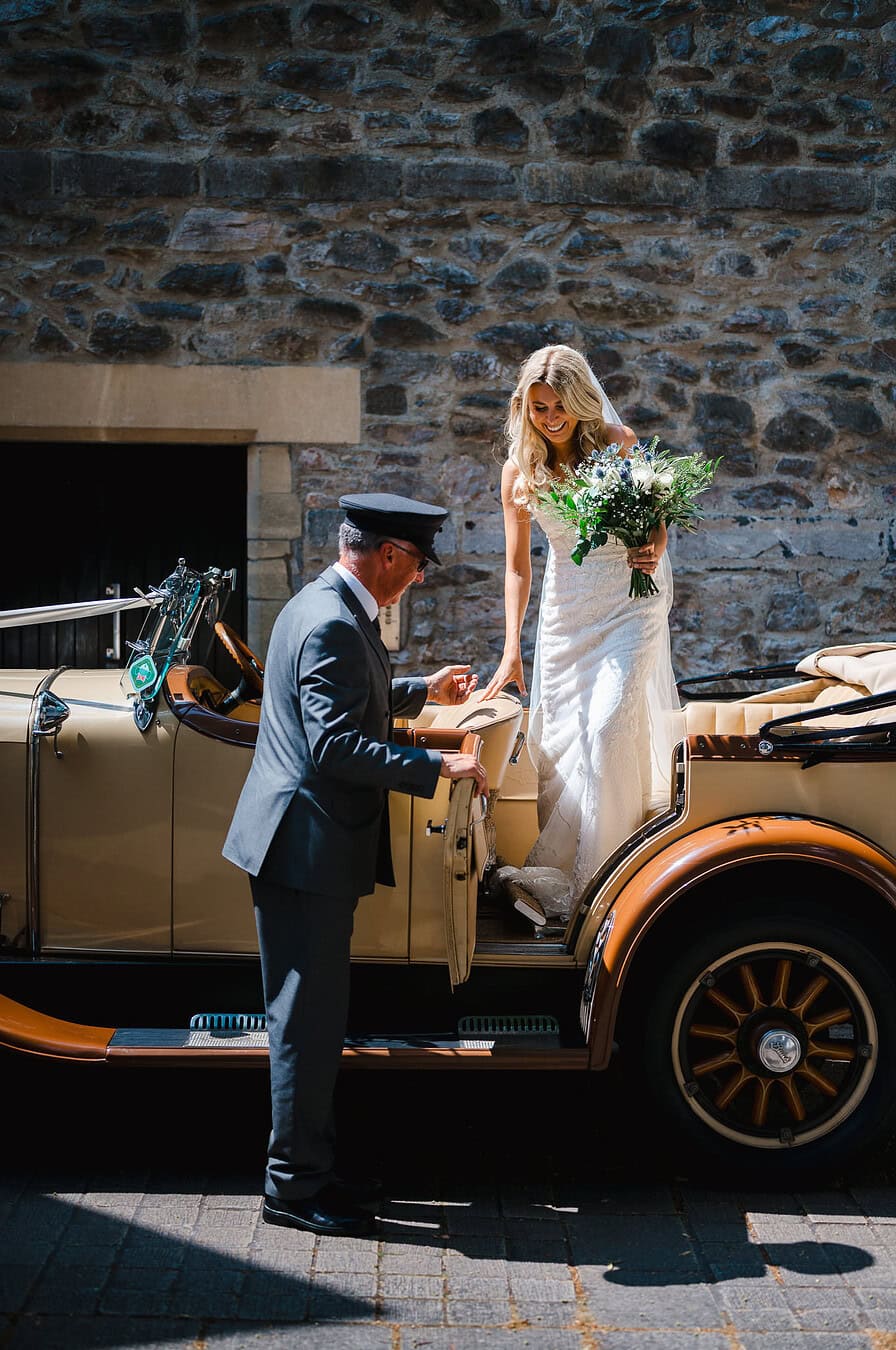The happy bride is about to make her wedding entrance as she steps out of her vintage wedding car at her wedding venue by Bournemouth Wedding Photographer, Jake Timms Photographer