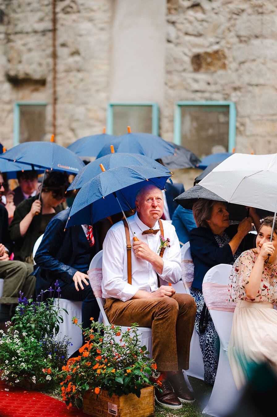 The rain starts to pour during their outdoor wedding as the father of the bride pulls a face from underneath the umbrella