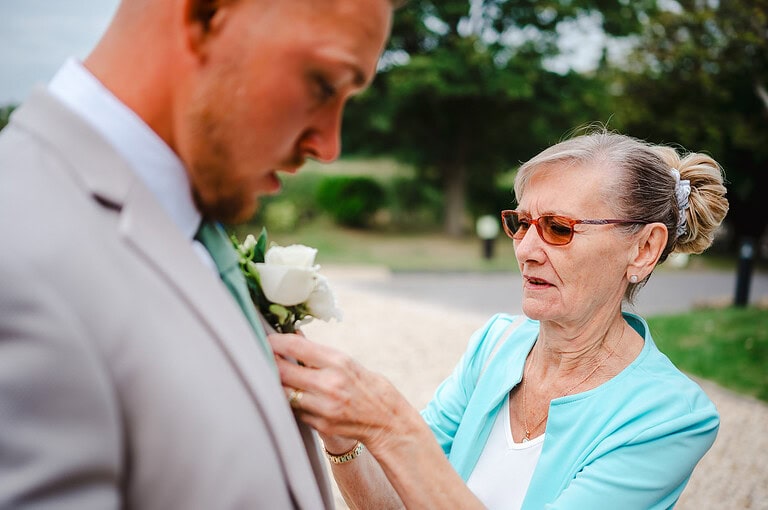 The groom has a helping hand as his buttonhole is placed on his suite jacket outside The Pear Tree By Jake Timms Photography