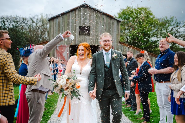 Wedding confetti pours over the happy, laughing couple as they walk away from their wedding ceremony barn -Documentary Wedding Photography by Jake Timms Photography