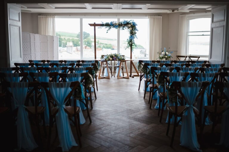 The wedding ceremony room with a beautiful flower archway at the end of the aisle surrounded by wooden chairs draped in a light blue cloth